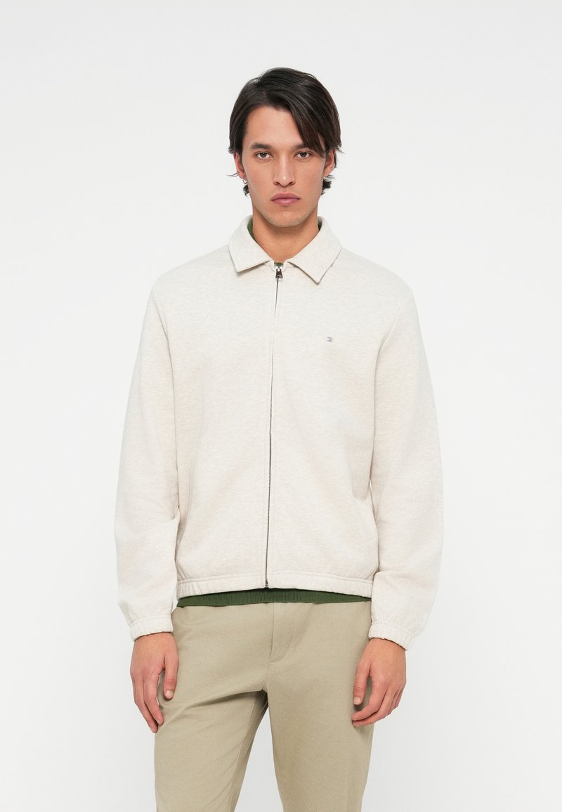 Jasno beżowa kurtka zapinana na zamek z kołnierzem, elastycznymi mankietami i pasem w talii. Zawiera małe logo na lewej piersi.