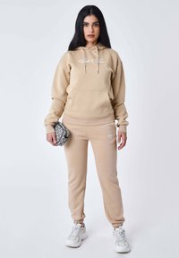 Sudadera beige y pantalones de chándal a juego, con un bolsillo frontal y detalle de logo, combinados con zapatillas blancas y una bolsa con estampado.