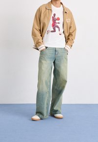 Persona con una chaqueta beige, camiseta blanca con un gráfico de un jugador de baloncesto, vaqueros claros holgados y zapatillas blancas sobre un suelo azul.