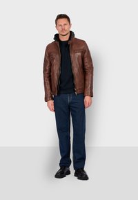 Bruine leren jack met een rits aan de voorkant en een fleece-gevoerde kraag, gecombineerd met een donkere trui en blauwe jeans, gedragen met zwarte schoenen.
