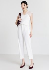 Donna che indossa un top con maniche in pizzo bianco, pantaloni bianchi su misura, tacchi slingback bordeaux e porta una borsa a tracolla bordeaux, in piedi davanti a uno sfondo semplice.