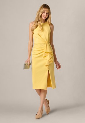 Kobieta w żółtej, midi sukience bez rękawów z falbaną i rozcięciem, trzymająca srebrną kopertówkę, ubrana w cieliste, szpiczaste szpilki, stojąca na szarym tle.