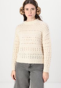Off-white, strikket genser med ribbestrikket hals og teksturert mønster med åpen maske detaljering. Kombinert med grå jeans.