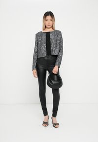 Fonkelende grijze pailletten cropjack, zwart getailleerd topje, zwarte leren broek, zwarte handtas en stippenhakken. Effen witte achtergrond.
