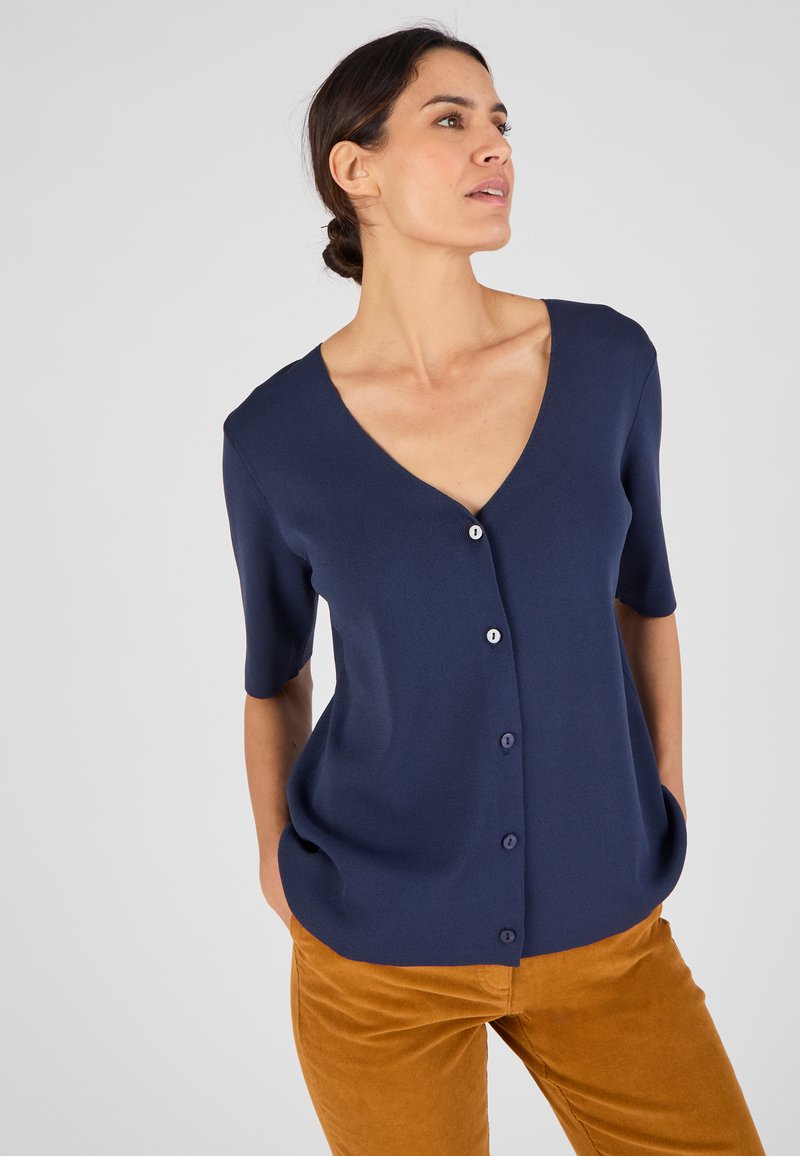 Cárdigan corto de manga corta en color marino con cuello en V y cierre de botones. Hecho de una tela texturizada, cuenta con dos bolsillos frontales y un corte relajado.