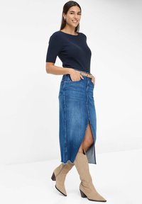 Denimrok met een hoge split, blauwe kleur en rafelige zoom; gecombineerd met een marineblauw ribbelshirt en tan knielange laarzen.
