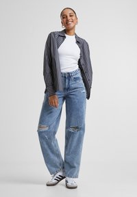 Gestreepte overhemd met lange mouwen, witte tanktop, versleten blauwe jeans met scheuren, en grijze sneakers met zwarte strepen.