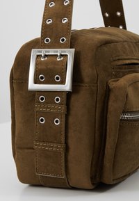 Sac en daim marron avec boucle argentée et œillets sur la sangle, avec une poche zippée latérale et des détails de couture visibles.