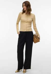 Femme portant un haut à manches longues beige boutonné, un pantalon noir à jambes larges, des escarpins à motif serpent, et tenant un sac à main marron sur un fond blanc.