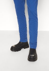 Mustat nahkasaappaat, joissa on paksu, teksturoitu pohja ja hopeiset yksityiskohdat, yhdistettyinä istuviin sinisiin housuihin.