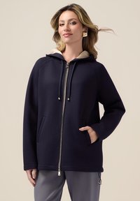 Felpa con zip di colore blu navy, con cappuccio foderato in beige, dotata di due tasche frontali, cordini tono su tono e dettaglio della zip metallica. Texture liscia.
