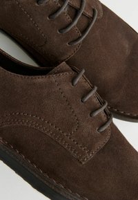 Zapatos de vestir de ante marrón con una textura suave, diseño con cordones y acentos cosidos en la parte superior. Punta redondeada y suela delgada de goma.