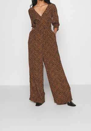Femme portant une combinaison pantalon marron et noire à larges jambes, avec un profond col en V et des manches trois-quarts, debout devant un fond blanc uni.