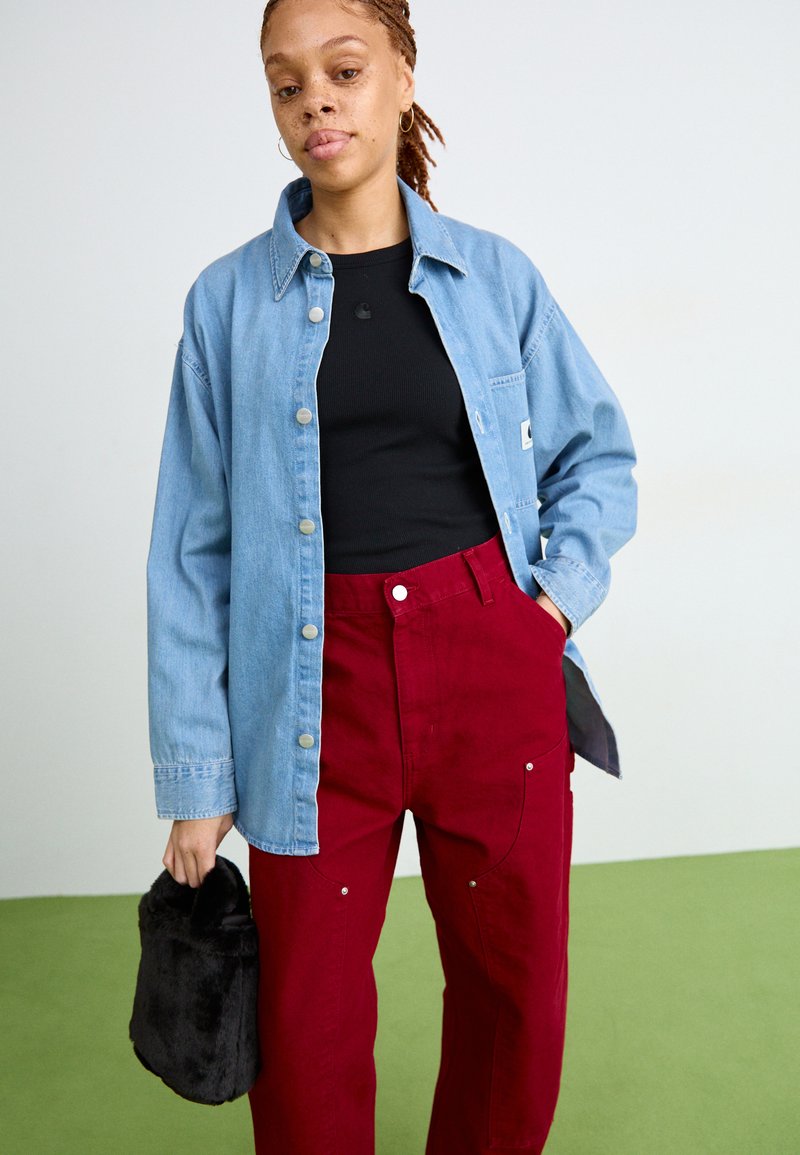 Lichtblauwe denim shirt met zilveren knopen, gecombineerd met hooggesneden rode broeken met zakken. Zwarte kortharige tas in de hand. Groene vloer.