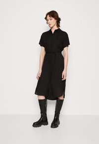 Abito nero a camicia con maniche corte, bottoni frontali e una cintura in vita. Abbinato a stivali neri al ginocchio. Tessuto liscio, vestibilità comoda.