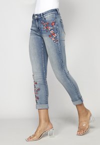 Hellblaue Jeans mit floralen Stickereien in Rot und Weiß an beiden Beinen. Aufgeschlagene Säume und eine taillierte Passform. Transparente Absatzsandalen.