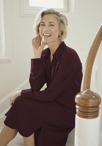 Robe bordeaux à boutons, à manches longues, en tissu texturé, assise sur un escalier. Accents en bois chaleureux en arrière-plan.