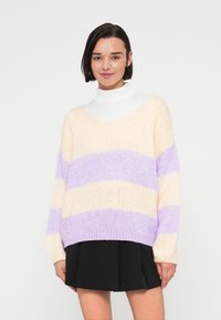 Pull en maille oversize crème avec des rayures horizontales lavande et violet clair. Doté d'un col en V et de poignets côtelés. Associé à une jupe noire.