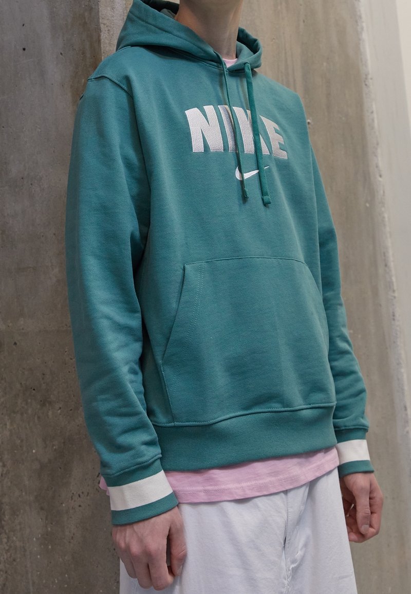 Tealfärgad hoodie med vitt "NIKE"-logo, dragsnören, känguruficka och ribbade muddar. Buren över en ljusrosa tröja, kombinerad med vita byxor.