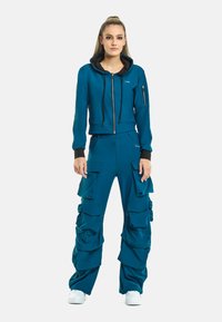 Teal cropped hoodie met zwarte voering, ritssluiting en een zak. Bijpassende teal cargobroek met meerdere zakken en een gerimpeld ontwerp. Witte sneakers.