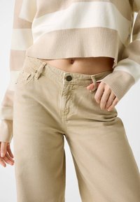 Beżowe spodnie o szerokich nogawkach z wysokim stanem, wyposażone w zapinanie na guzik, fason z pięcioma kieszeniami oraz gładką, teksturowaną bawełnianą tkaninę.