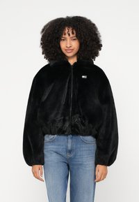 Γυναίκα με κατσαρά μαλλιά που φοράει μαύρο χνουδωτό μπουφάν με φερμουάρ και μπλε τζιν, στέκεται μπροστά σε απλό λευκό φόντο.