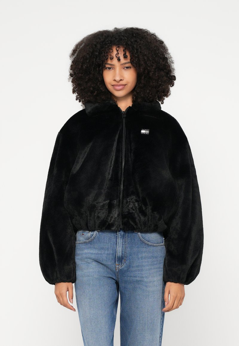 Γυναίκα με κατσαρά μαλλιά που φοράει μαύρο χνουδωτό μπουφάν με φερμουάρ και μπλε τζιν, στέκεται μπροστά σε απλό λευκό φόντο.