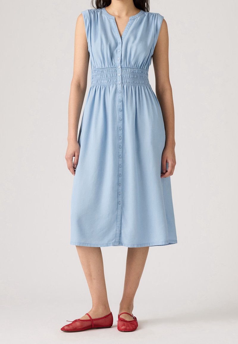 Robe midi bleu clair sans manches avec boutons sur le devant et taille smockée, associée à des ballerines en maille rouge sur une femme debout.
