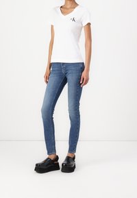 Camiseta blanca de cuello en V con el logo de CK, jeans ajustados azules y zuecos de plataforma negros. Tejido suave, diseño ceñido y mangas relajadas.