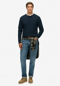 Jonge man in een marineblauwe longsleeve henley, blauwe jeans, beige schoenen, een geruit shirt om zijn taille geknoopt, en een polshorloge, staand tegen een effen achtergrond.