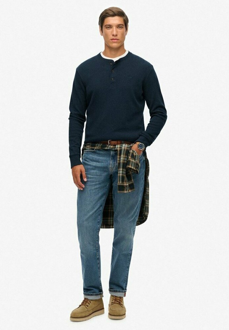 Jonge man in een marineblauwe longsleeve henley, blauwe jeans, beige schoenen, een geruit shirt om zijn taille geknoopt, en een polshorloge, staand tegen een effen achtergrond.