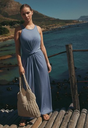 Femeie în rochie albastră fără mâneci, ținând o geantă împletită, stând pe un ponton de lemn lângă mare, cu o coastă muntoasă în fundal.