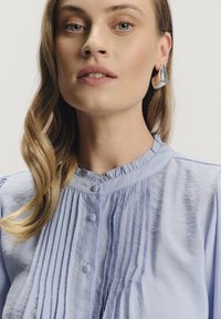 Blusa plissettata azzurro chiaro con colletto arricciato e chiusura con bottoni. Capelli acconciati in onde morbide, indossando un orecchino a goccia in argento.