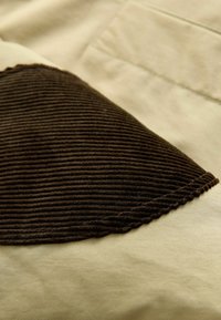 Tessuto di cotone con una superficie liscia color crema, caratterizzato da una toppa ribattuta marrone scuro con un bordo ondulato nell'angolo in alto a sinistra.