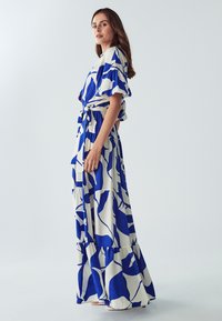 Sininen ja valkoinen kuvioitu maksimekko, jossa on lyhyet röyhelösateet, vyötärö on kiristetty ja hameen muotoilu on kerroksellinen. Sujuva kangas, tyylikäs liikkuvuus.