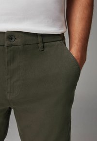Pantalones de algodón verde oscuro con un bolsillo frontal, lazos para cinturón y un botón visible. La textura parece suave con detalles de costura reforzada.