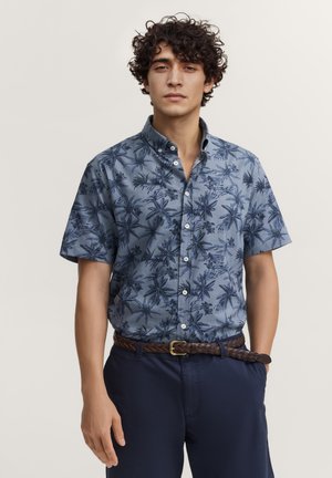 Junger Mann mit lockigem Haar, der ein blaues, kurzärmliges Hemd mit Blumenmuster, marineblaue Hose und einen geflochtenen braunen Gürtel trägt, steht vor einem schlichten Hintergrund.