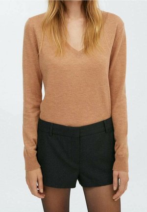 Frau trägt einen kamelfarbenen V-Ausschnitt-Pullover, schwarze maßgeschneiderte Shorts und transparente schwarze Strumpfhosen, steht vor einem schlichten Hintergrund.