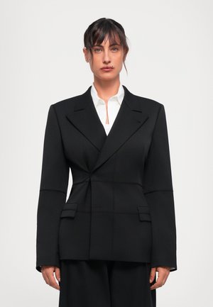 Femme aux cheveux foncés portant un blazer noir sur une chemise blanche à boutons, faisant face à l'avant contre un fond gris clair.