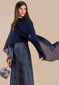 Chal navy de gasa sobre un vestido azul de manga larga con estampado floral. La modelo sostiene un bolso plateado texturizado, con un fondo beige suave.