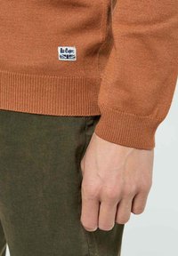 Pull en maille marron avec des poignets et un ourlet à côtes. Présente un tissu texturé et un petit patch logo blanc près du bas à gauche.