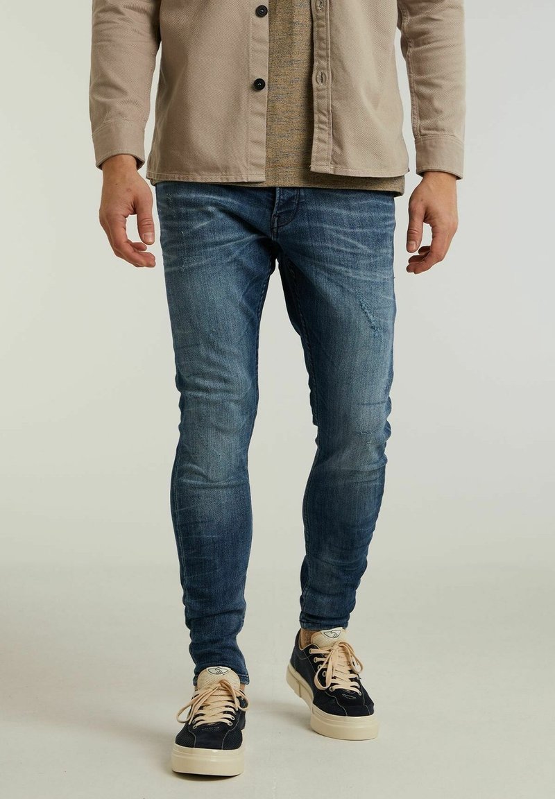 Homme portant un jean skinny bleu délavé, des baskets noires et beiges, et une veste beige, debout sur un fond clair.