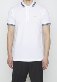 Homme portant un polo blanc à manches courtes avec un col et des bords de manches rayés bleu et noir, assorti à un pantalon noir.
