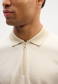 Beige polo skjorte laget av teksturert stoff med glidelåskrage, som har et subtilt geometrisk mønster. Nærbilde av nakken og brystet.
