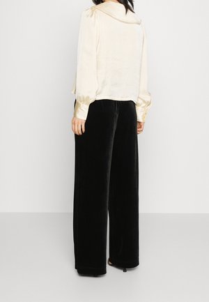 Nainen pukeutuneena kermaiseen satiinipaitaan, jossa on pitkät hihat, ja mustiin leveälahkeisiin samettihousuihin, seisoo selkä kameraan päin yksinkertaista taustaa vasten.