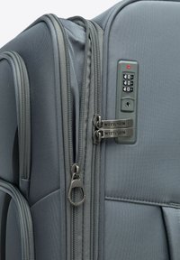 Grijze koffer met textuurstof, duale ritsen, combinatieslot en merkrits. Compact en gestructureerd ontwerp voor reizen.