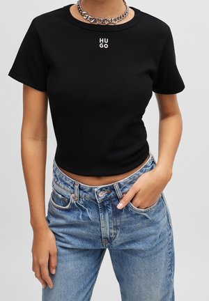 Osoba ubrana w czarny krótki T-shirt z białym napisem "HUGO" oraz jasnoniebieskie jeansy z wysokim stanem, uzupełniona srebrnym naszyjnikiem z łańcuszka.