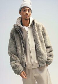 Jachetă din blană sintetică de culoare gri deschis, cu croială supradimensionată. Glugă atașată cu textură moale, purtată peste un hanorac bej cu fermoar și pantaloni de trening asortați.