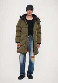 Olive green puffer coat with a hood, zip closure, and side pockets, paired with distressed blue jeans featuring patchwork details and black shoes.