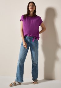 Top viola a maniche corte con nodo laterale, abbinato a jeans in denim a gamba larga di colore blu chiaro. La persona indossa sandali argentati.